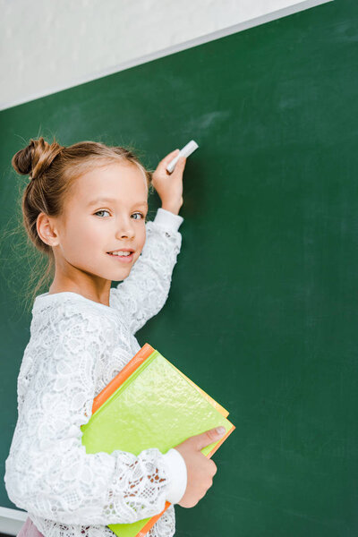 happy schoolkid holding chalk and books near green chalkboard 