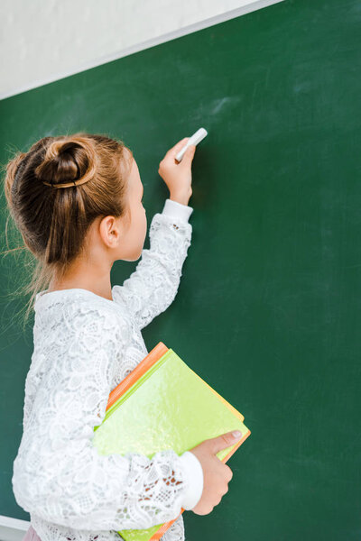 cute schoolkid holding chalk and books near green chalkboard 