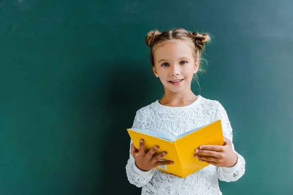 Happy Schoolgirl Tersenyum Sambil Berdiri Dengan Buku Berwarna Hijau — Stok Foto