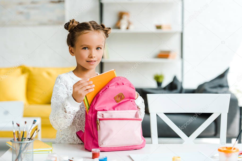 happy child holding book near pink backpack at home 