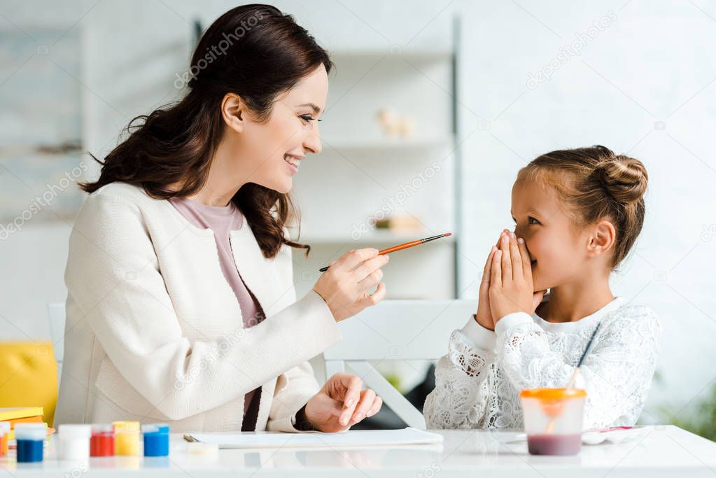 attractive mother holding paintbrush near happy daughter covering face