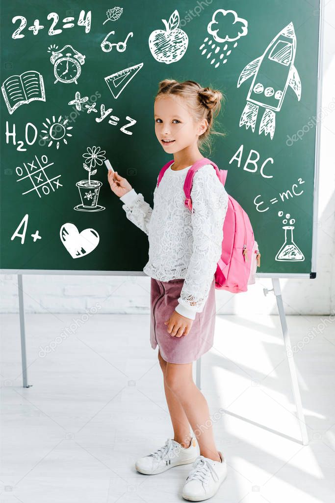 happy kid standing with backpack and holding chalk near drawing on green chalkboard 