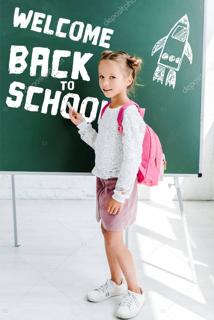 cute schoolgirl standing with backpack and holding chalk near welcome back to school lettering on green chalkboard 