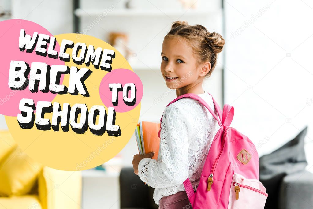 smiling schoolchild holding books while standing with backpack near welcome back to school letters 