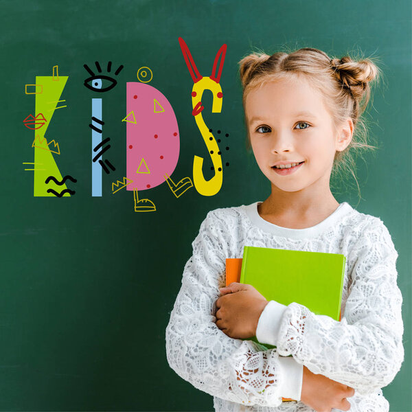 happy schoolgirl smiling while standing with books near kids lettering on green 
