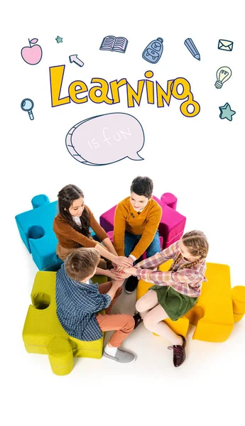 high angle view of kids sitting on jigsaw puzzle poufs and stacking hands near learning  is fun letters on white