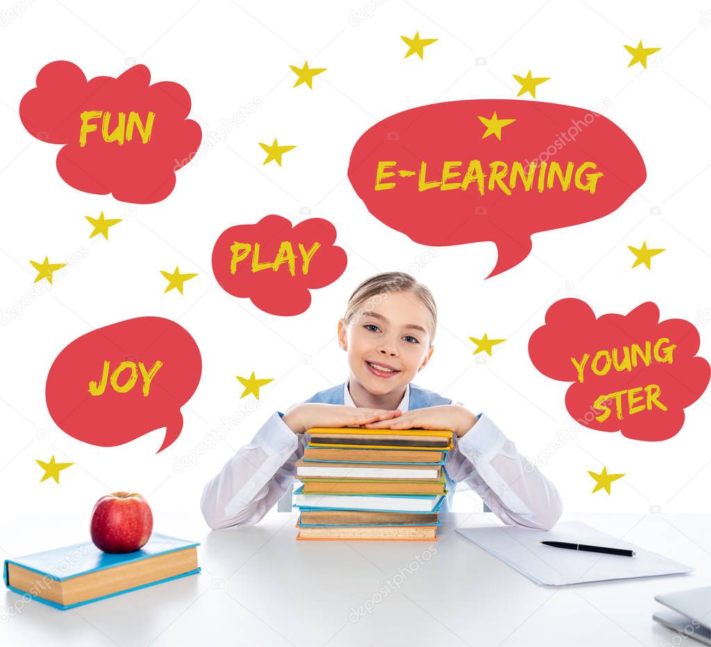 smiling schoolgirl sitting at desk with books and looking at camera near speech bubbles with letters on white 