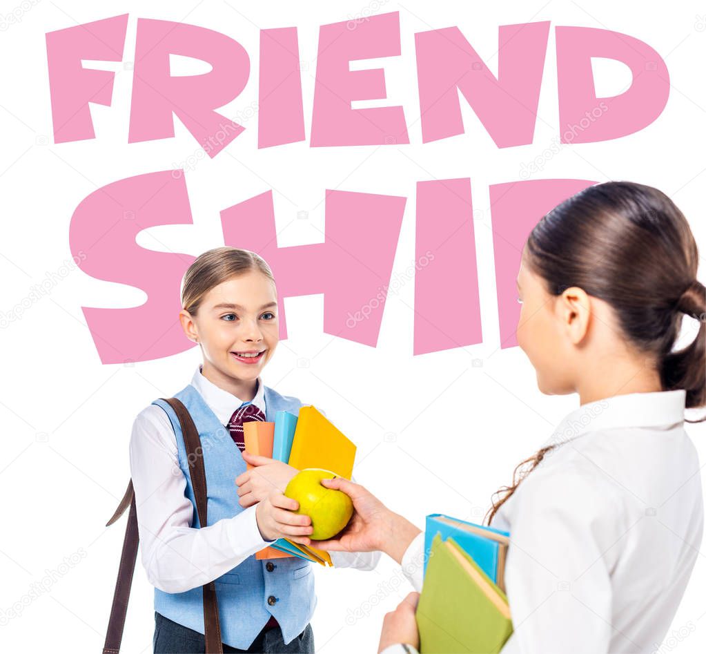 schoolgirls in formal wear with books looking at each other and sharing apple near friendship letters on white 