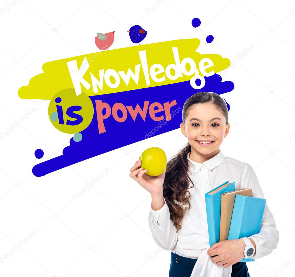 smiling schoolgirl holding apple and books while looking at camera near knowledge is power letters on white 