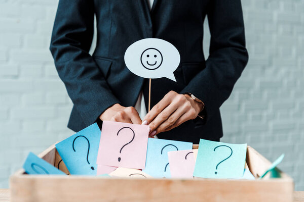 KYIV, UKRAINE - AUGUST 5, 2019: cropped view of businesswoman holding stick with smiley face near box with question marks 