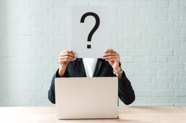 cropped view of woman covering face while holding board with question mark near brick wall and laptop clipart