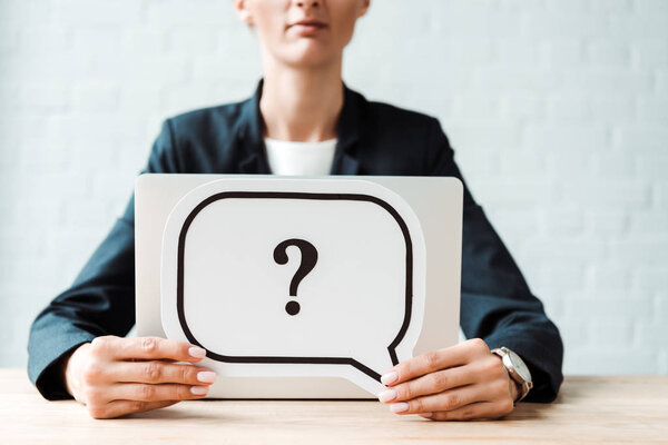 cropped view of woman holding black speech bubble with question mark near desk 