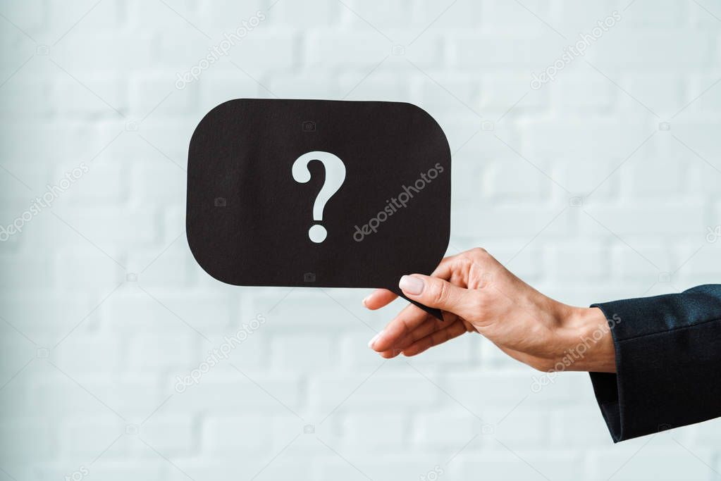 cropped view of woman holding black speech bubble with question mark near white brick wall 
