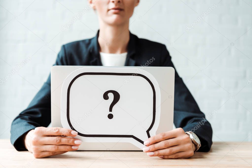 cropped view of woman holding black speech bubble with question mark near desk 