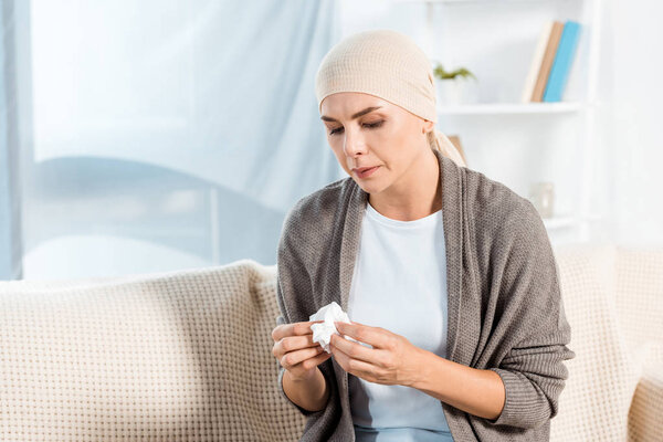 sick woman with head scarf holding tissue while sitting on sofa 