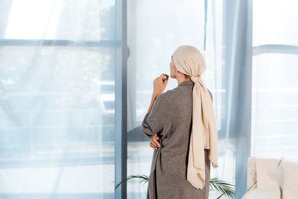 sick woman with head scarf standing in living room near windows 