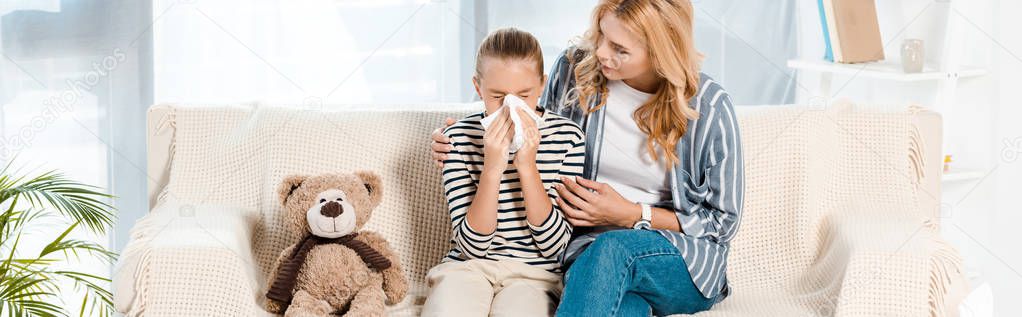 panoramic shot of woman sitting near daughter sneezing in tissue near teddy bear 