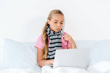 sick kid holding digital thermometer in mouth and looking at laptop  clipart