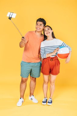 yakışıklı asyalı adam sarı arka plan üzerinde plaj topu tutan kız arkadaşı yanında selfie sopa ile akıllı telefonda selfie alarak