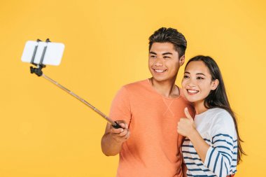yakışıklı asyalı adam kız arkadaşı yanında selfie sopa ile akıllı telefonda selfie alarak sarı izole başparmak gösteren