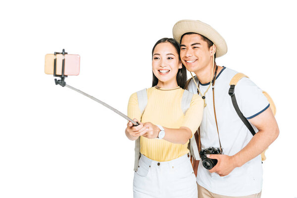 two cheerful asian tourists taking selfie on smartphone with selfie stick isolated on white