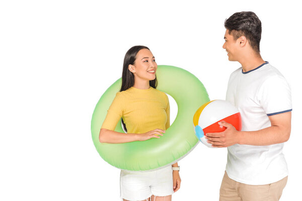 handsome asian man holding beach ball while looking at cheerful girlfriend with swim ring isolated on white