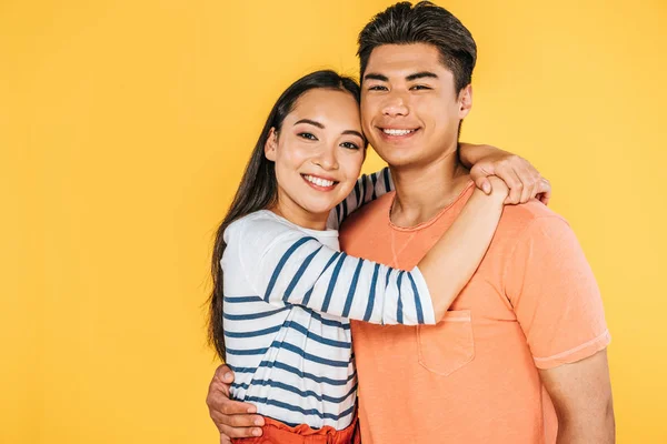 happy young asian couple embracing while smiling at camera isolated on yellow