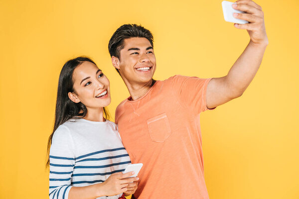 happy asian man taking selfie on smartphone with beautiful girlfriend isolated on yellow