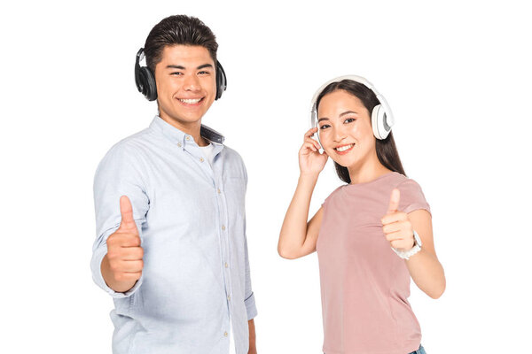 smiling asian man and woman showing thumbs up while listening music in headphone and looking at camera isolated on white