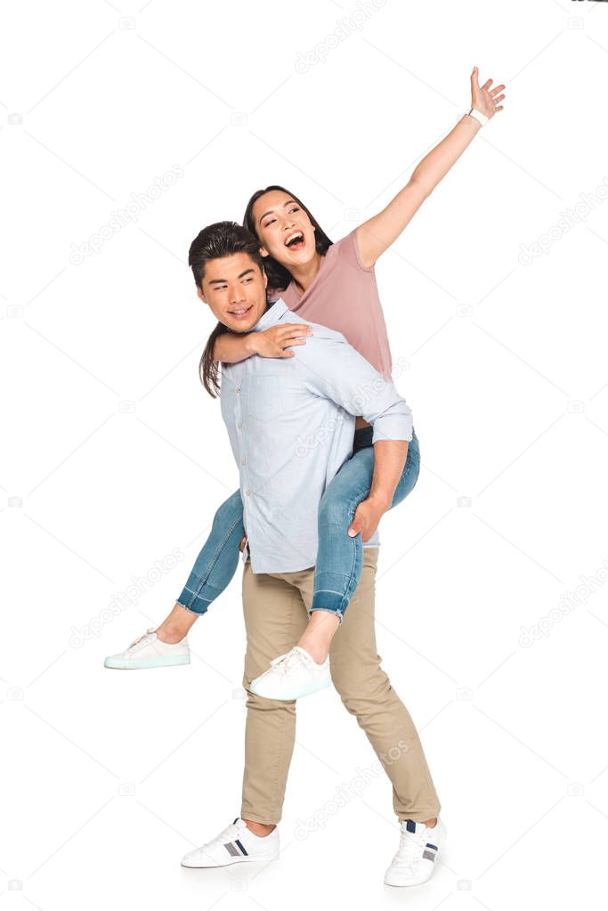 cheerful asian woman waving hand while piggybacking on happy boyfriend on white background