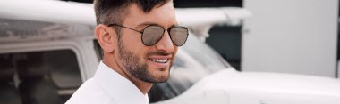 panoramic shot of smiling bearded pilot in sunglasses standing near plane clipart