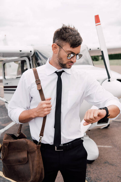 bearded pilot in sunglasses standing near plane and looking at wristwatch