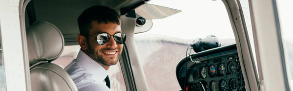 panoramic shot of smiling handsome pilot in sunglasses looking at camera in plane