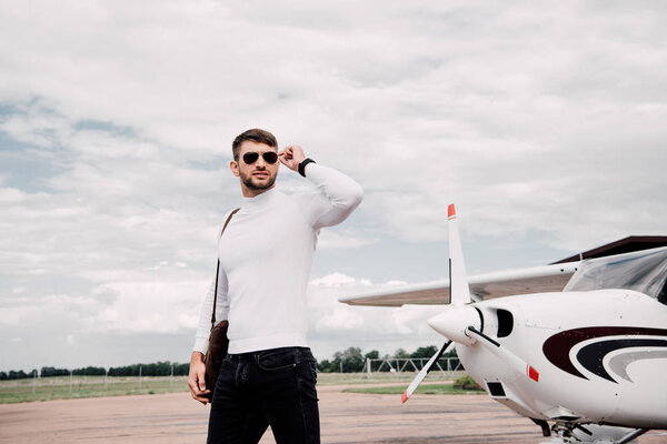 man in sunglasses with bag standing near plane under cloudy sky