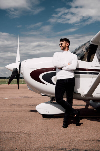 full length view of man in sunglasses standing with crossed arms near plane in sunny day