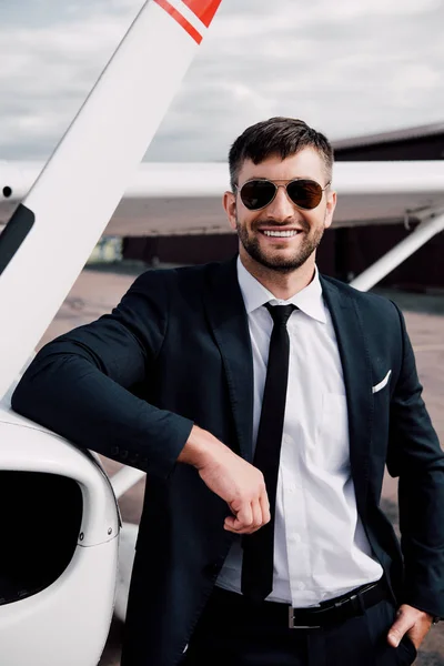 飛行機の近くにポケットに手を入れて立っているフォーマルな服を着て笑顔のビジネスマン — ストック写真