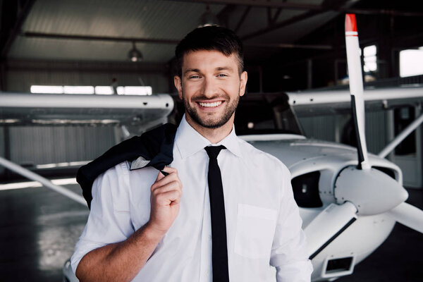 smiling pilot in formal wear standing near plane and looking at camera