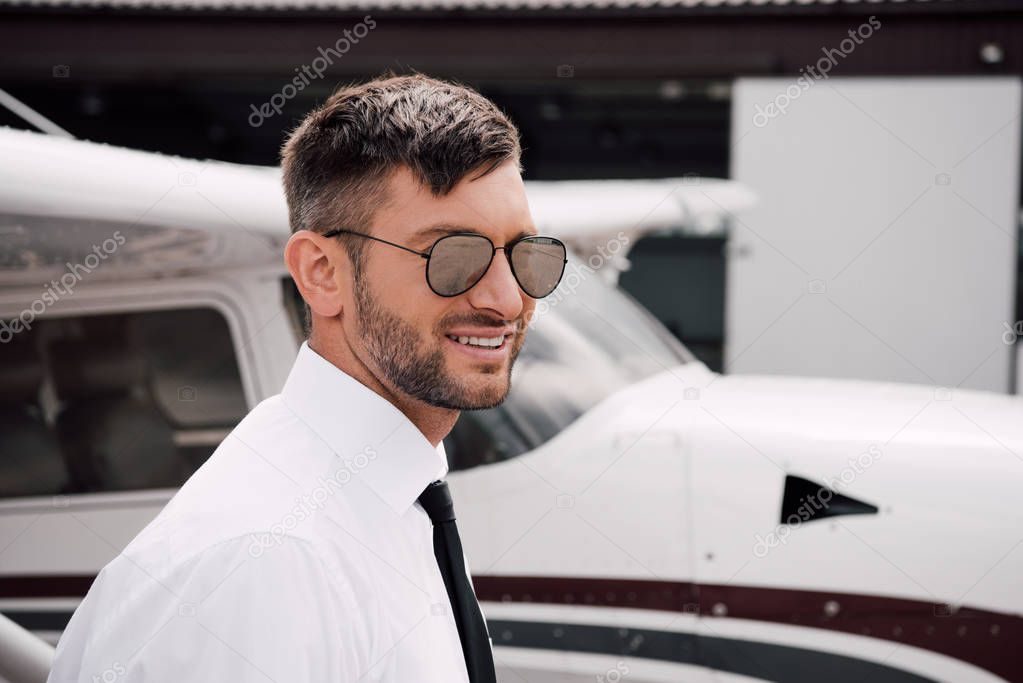 bearded pilot in formal wear and sunglasses smiling near plane