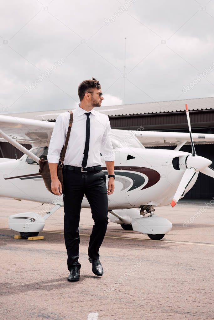 full length view of confident pilot with bag standing near plane under cloudy sky