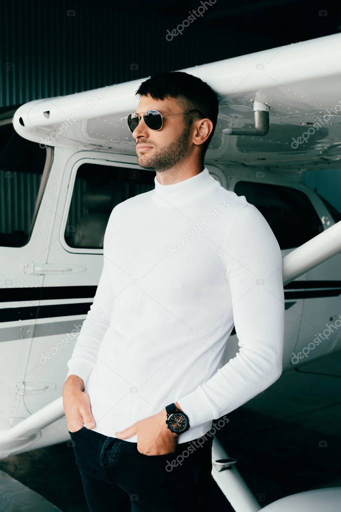 handsome young man in casual outfit standing with hands in pockets near plane and looking away