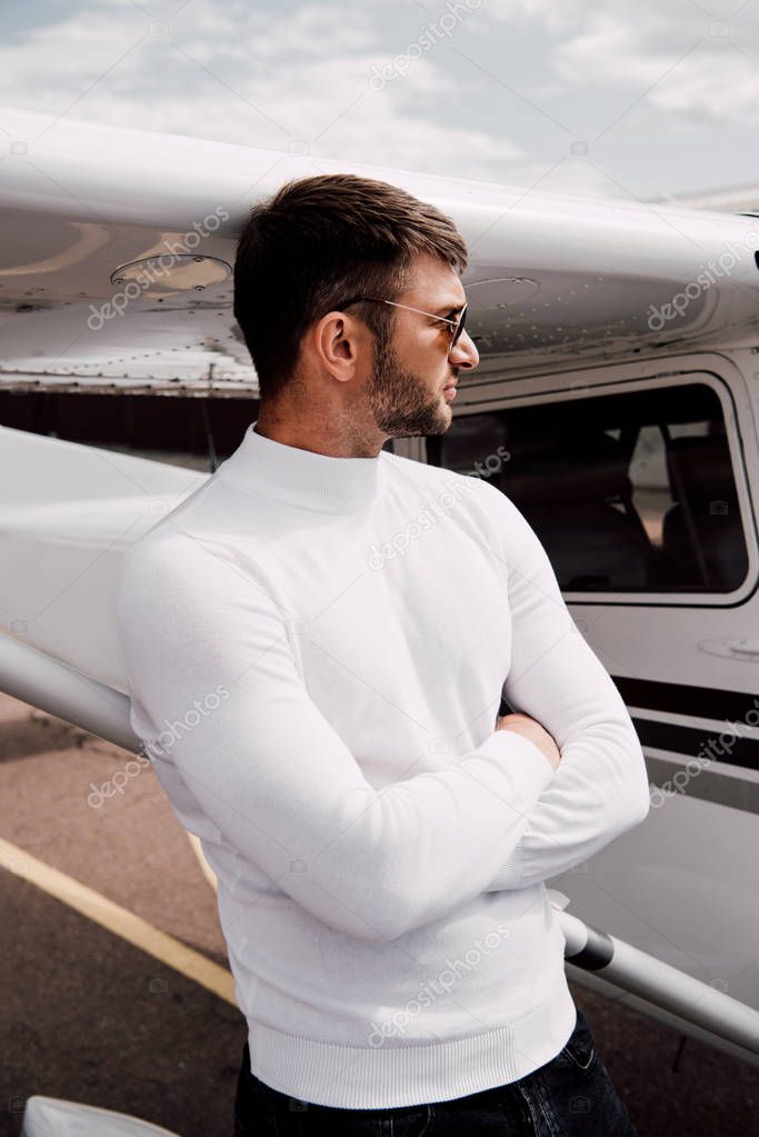 pensive bearded man in sunglasses standing with crossed arms near plane
