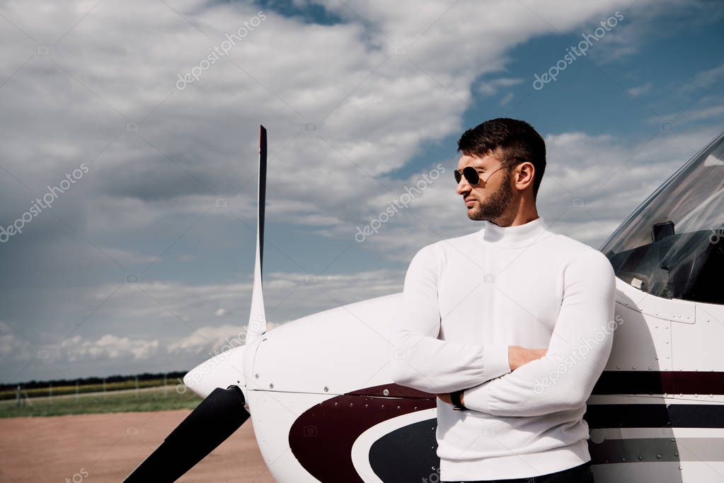 man in sunglasses standing with crossed arms near plane in sunny day