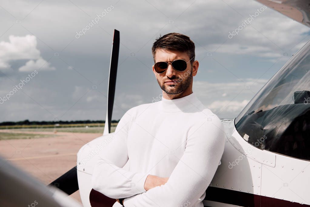 man in sunglasses standing with crossed arms near plane in sunny day