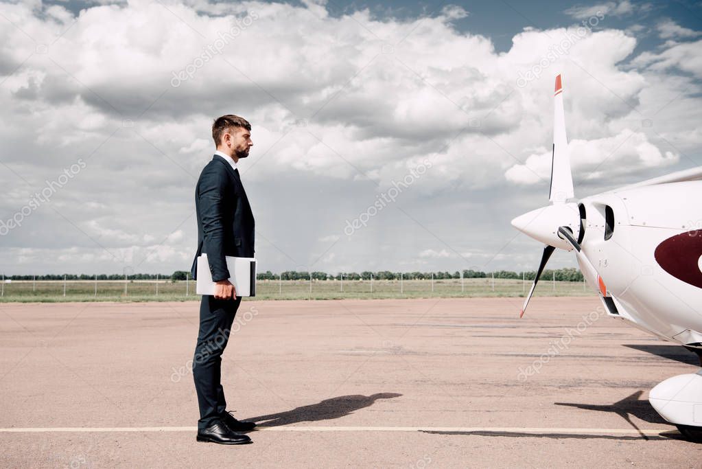 side view of businessman in formal wear holding laptop and folder near plane in sunny day