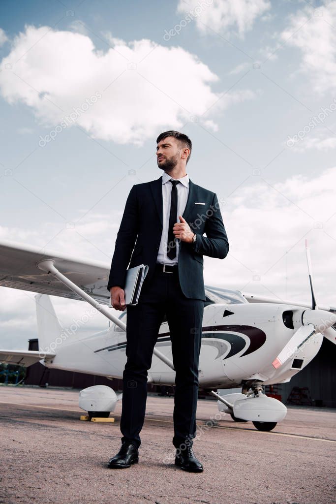 full length view of businessman in formal wear holding laptop near plane in sunny day