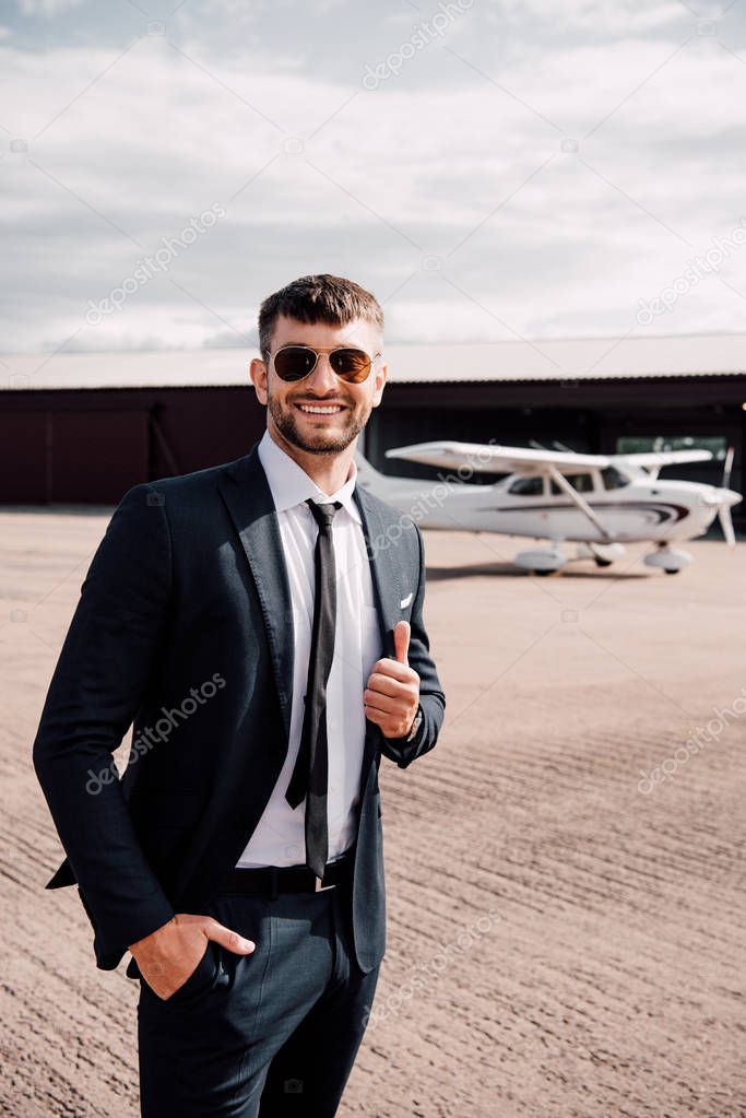 businessman in formal wear standing with hand in pocket in front of plane and showing thumb up