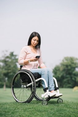 pretty disabled woman in wheelchair using smartphone while resting in park clipart