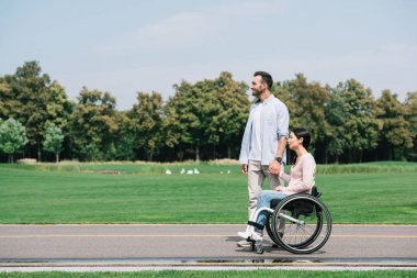 handsome young man holding hands with disabled girlfriend while walking in park together clipart
