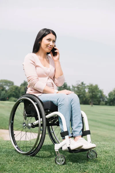 happy disabled woman in wheelchair talking on smartphone while resting in park