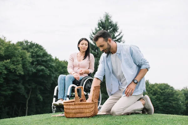 公園で障害のあるガールフレンドの近くの芝生に座っている間にストローバスケットを開くハンサムな男 — ストック写真